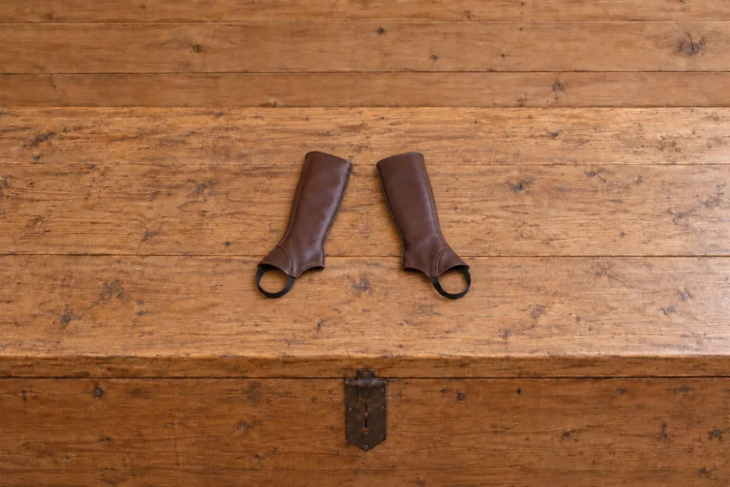 Bottes & Boots Une paire de mini-chaps en cuir marron posée sur une malle en bois devant un mur de planches nu.